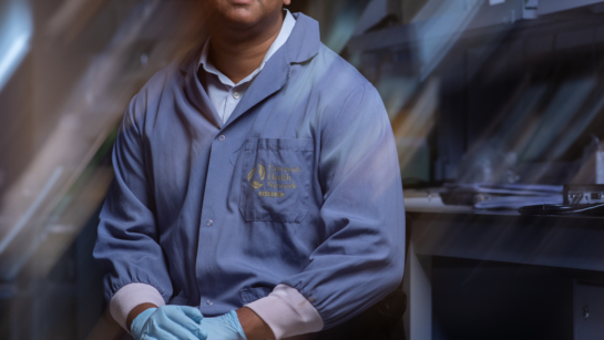 Dr. Karun Singh in the lab with lab coat and gloves sitting on a stool. Background around him is blurred for an artistic look so only he is in focus.