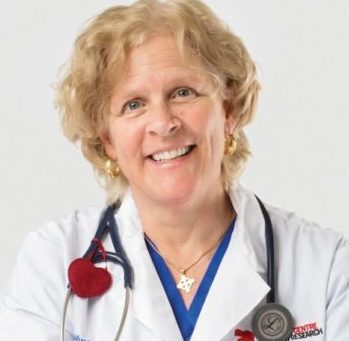 Photo of Dr. Heather ross sitting at a plain white table, she is wearing a lab coat, smiling towards the camera and holding an iPhone in her right hand.