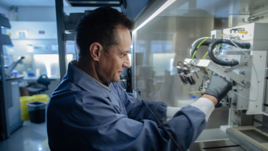 Dr. Michael Reber in the lab working with a machine that is behind a glass case. He is wearing black latex gloves and a lab coat.