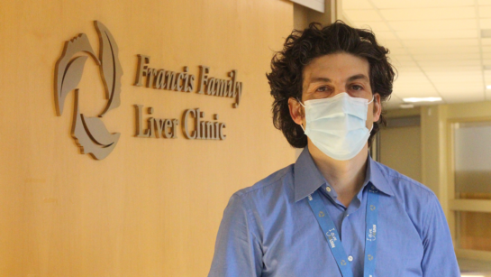 Photo of Dr. Jordan Feld standing in front of the Francis Family Liver Clinic sign. He is wearing a face mask, blue button-up shirt and has a lanyard on.