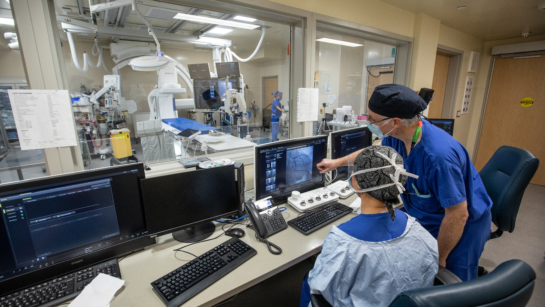 Photo of two technicians in scrubs looking at a computer. The computer is in a room with a glass wall that can see into the new Cath Lab filled with medical equipment.