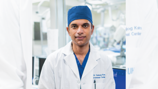 Close up photo of Dr. Sanjog Kalra in hospital. He is sitting against a desk and smiling, wearing lab coat and scrub cap.