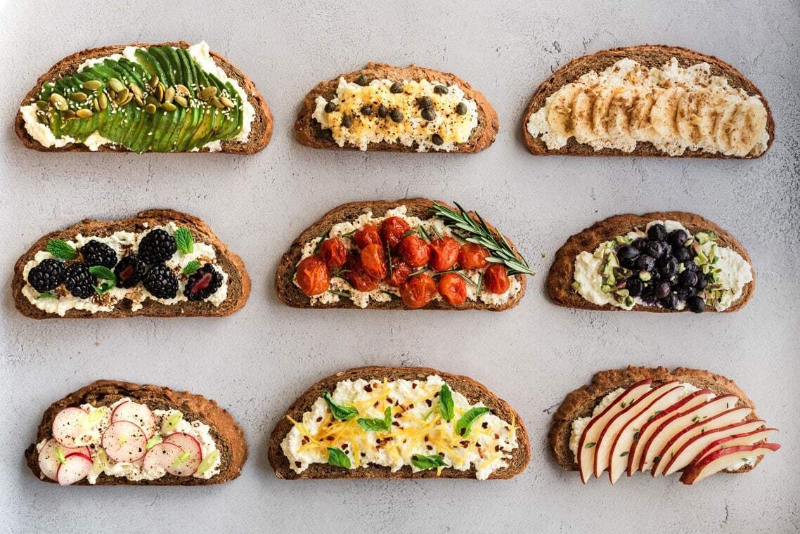 Flat lay photo of nine different toasts with various colourful toppings. Plain grey background.