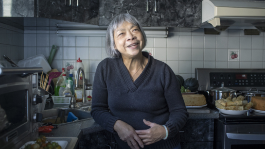 Photo of an eldery woman standing in her kitchen and smiling at the camera