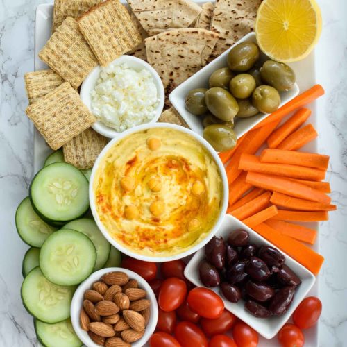 Flat lay photo of a hummus platter with hummus in the middle, vegetables around the sides, olives in smaller bowls on the platter, almonds, crackers and an additional dip. Half a lemon on the top right of the platter and the platter is sitting on a marble countertop. 