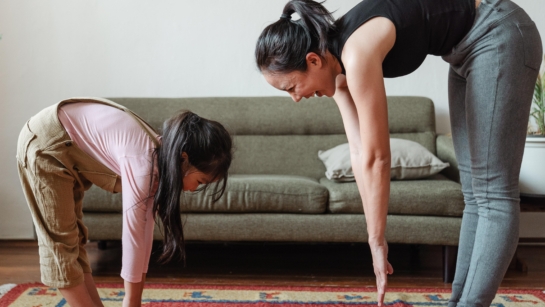 Landscape photo of a mother and daughter stretching (trying to touch their toes) together in the living room.