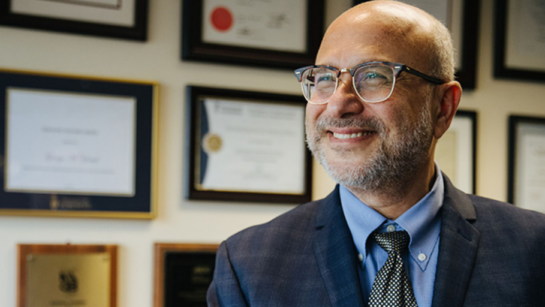 Dr. George Yousef smiles while wearing a blue suit and glasses in front of a wall of awards
