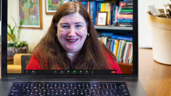 Dr. Susan Abbey appears on a laptop during a live stream. The laptop is on a wooden desk with a plant beside in the background.