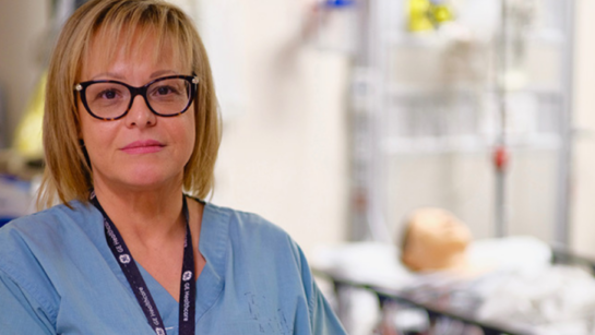 UHN's Maggie Dilling poses in her baby blue scrubs with a training simulation in the background.