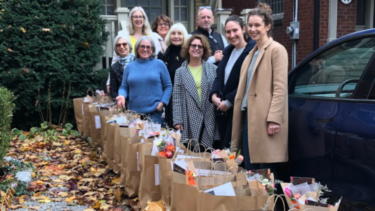 A group of volunteers stand in a drive way on a autumn day with rows of paper bags filled with food for delivery