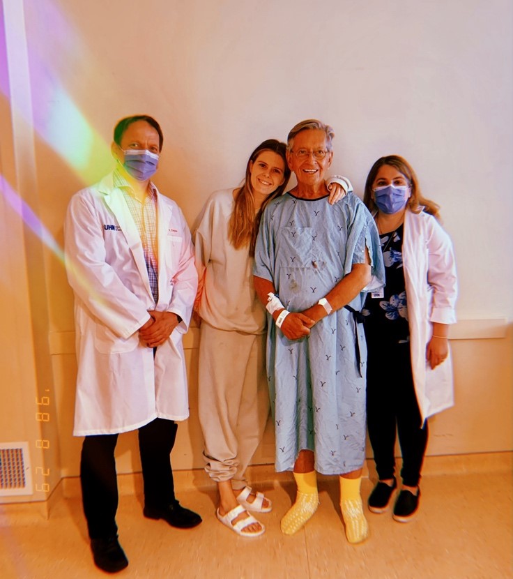 Meredith and her dad taking a photo together at UHN in a hallway while her dad is wearing a hospital gown.