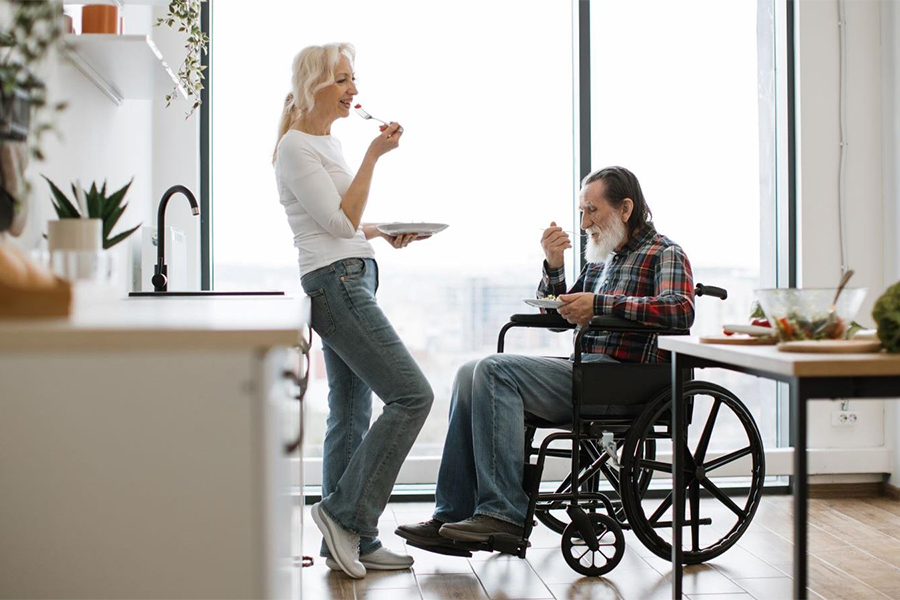 A photo of a woman standing and eating while leaning against a kitchen counter. A man is seated in a wheelchair next to her eating. A bright window is in the background with lots of light shining in.