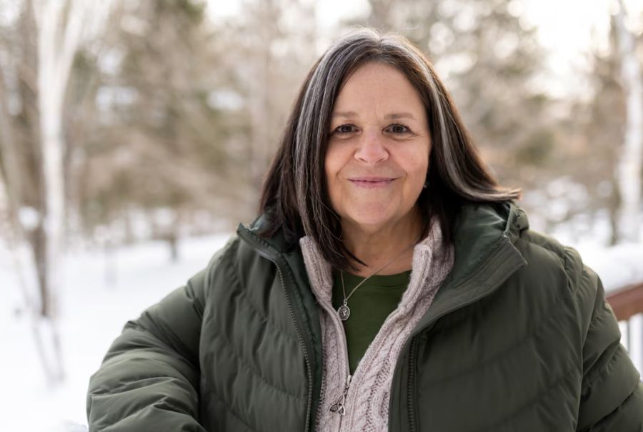 Kim Rose smiling in a winter coat while on a deck with a forest in the background.