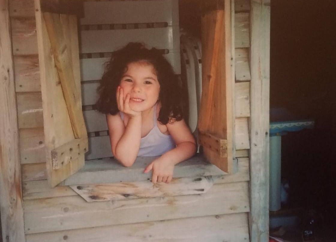 Macenzie Robelo posting in a wood fort with her hand on her chin and two split windows open and her leaning on it looking outward.