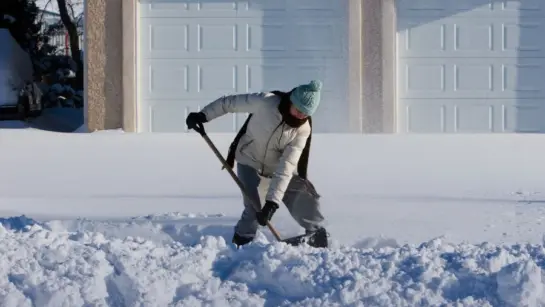 A person shovelling snow on a bright sunny day. There are two garage doors behind them as they are bent down with several inches of snow.