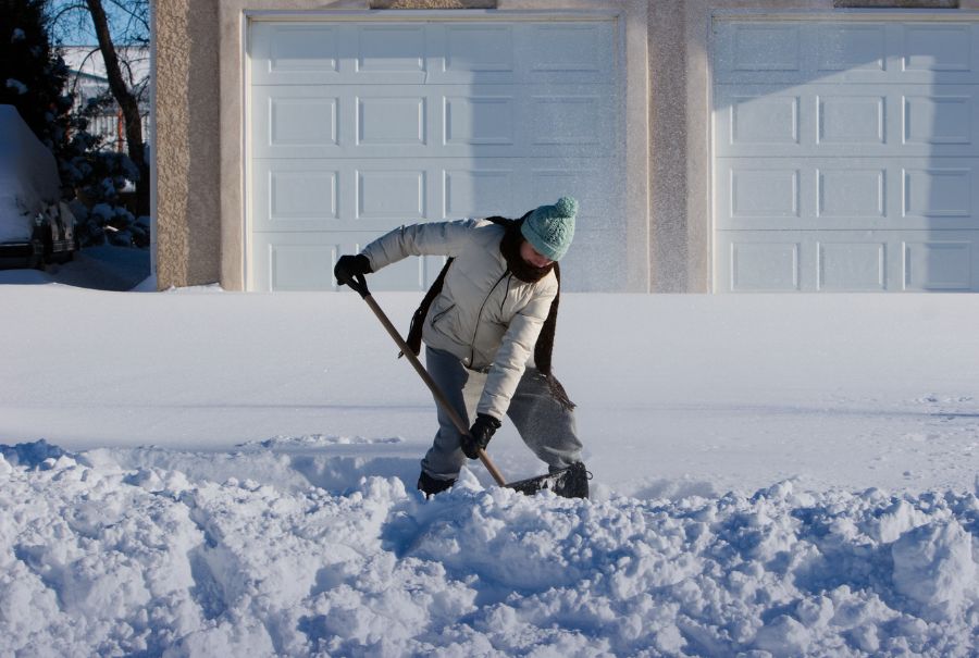 A person shovelling snow on a bright sunny day. There are two garage doors behind them as they are bent down with several inches of snow.