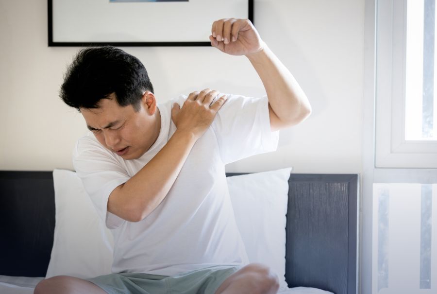 A man grabbing his shoulder and raising his arm while appearing to be in pain/discomfort. He's sitting on the edge of his bed.