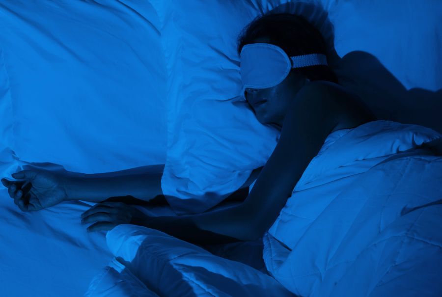 A person sleeping in bed with a sleep mask on. It's dark with a light blue light being shown in order to see the person laying in bed. 