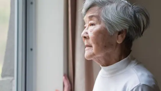 An older lady looking outside of her window as the light highlights her face.
