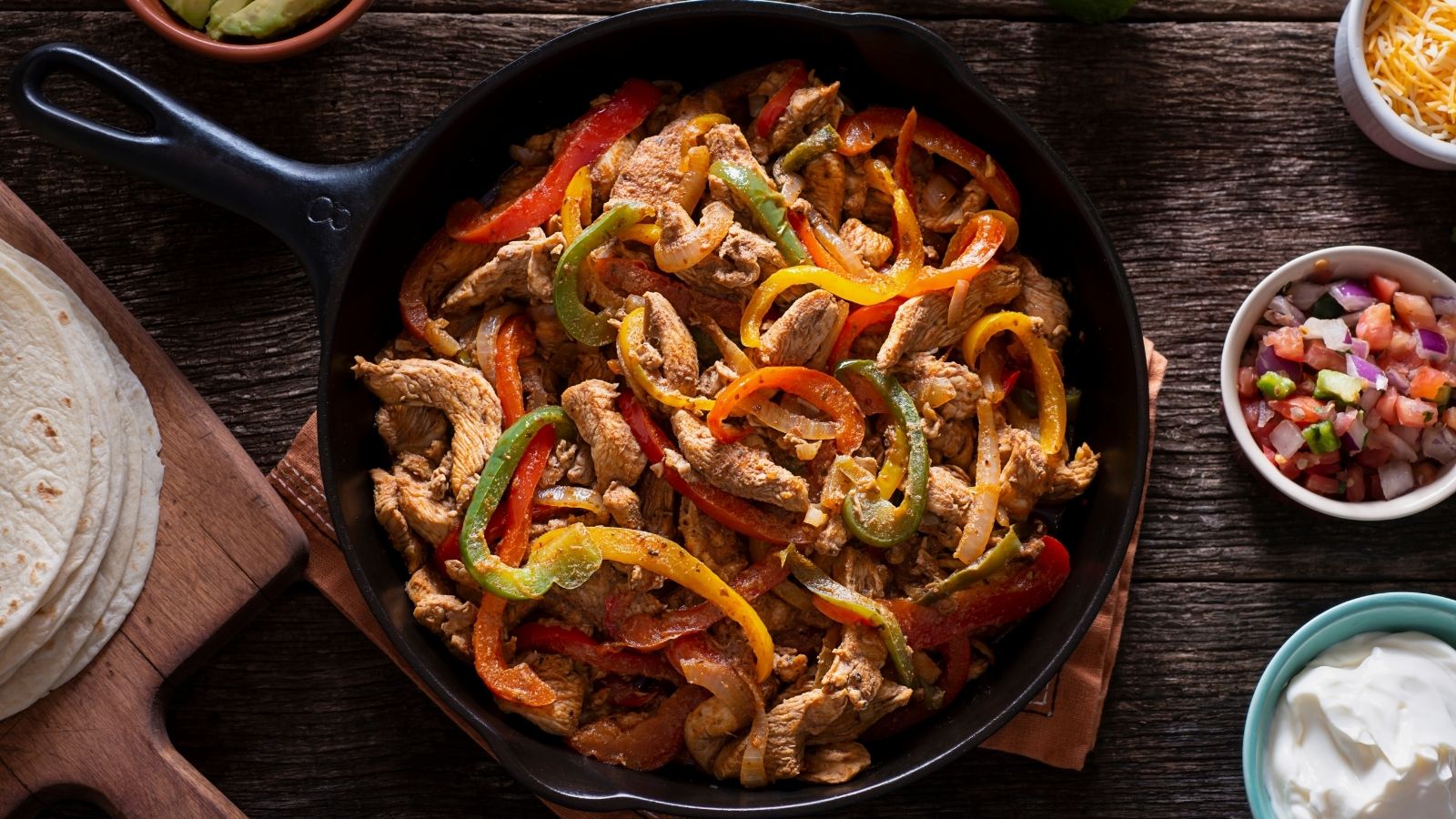 Chicken fajitas on a table with bell peppers mixed in. On the table are: tortillas, sour cream and cheese.
