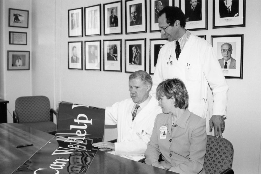 In a black and white photo, Dr. Michael Baker is standing in behind two people with a new sign being shown on a table.