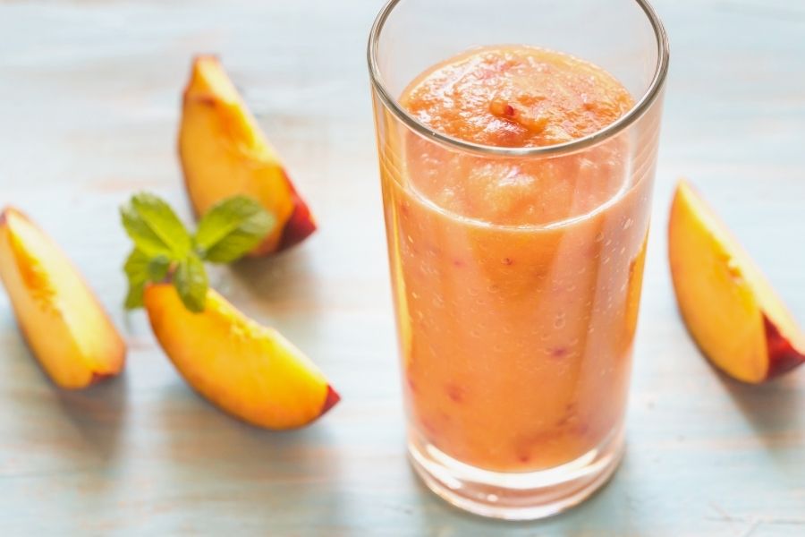 A peach smoothie in a glass on a table with four slices of peaches surrounding it.