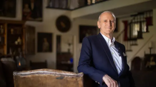 Donald K. Johnson standing in a room wearing a suite with his arm resting on a chair.