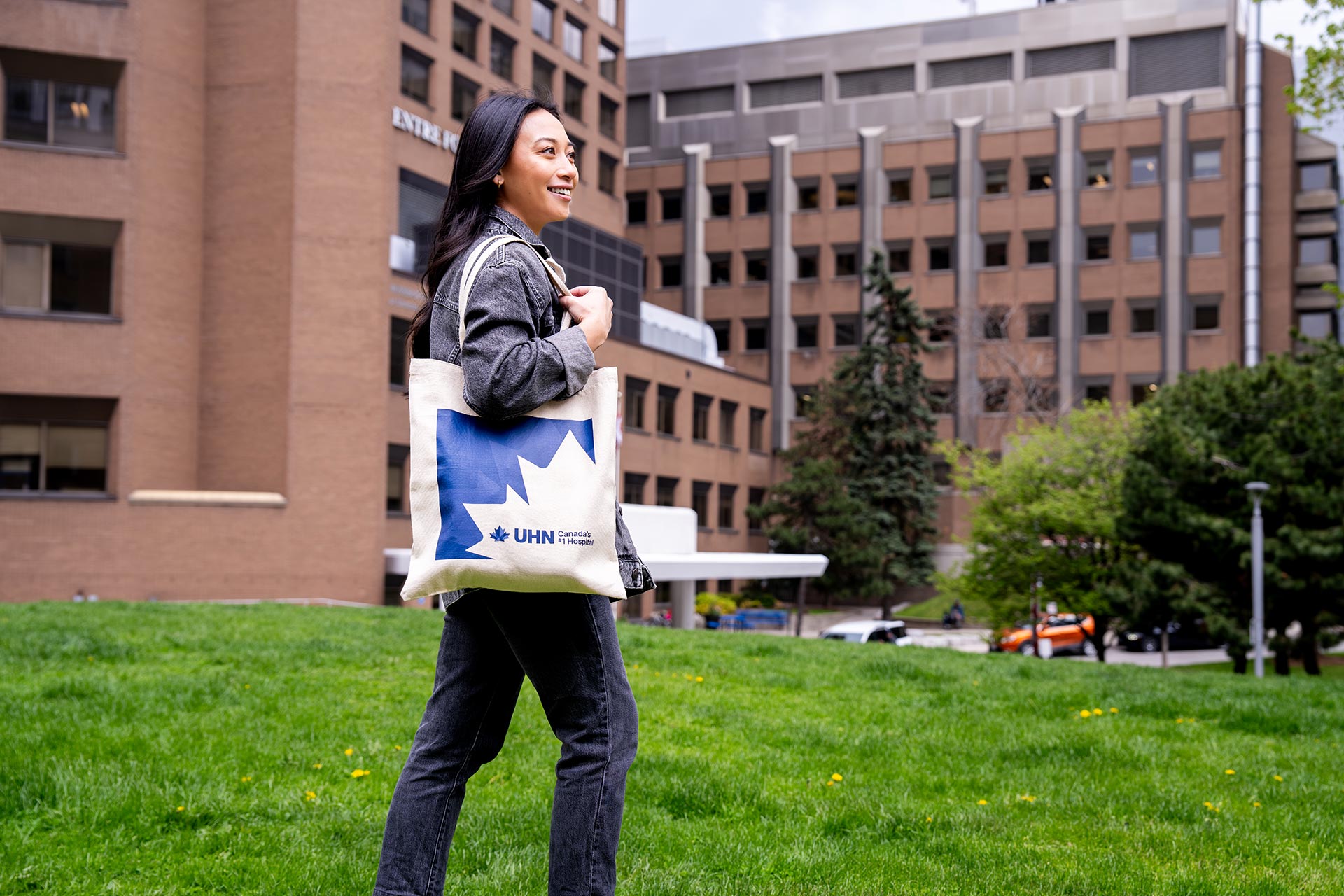 A woman stands outside a hospital building, smiling and carrying a bag with the UHN (University Health Network) logo.