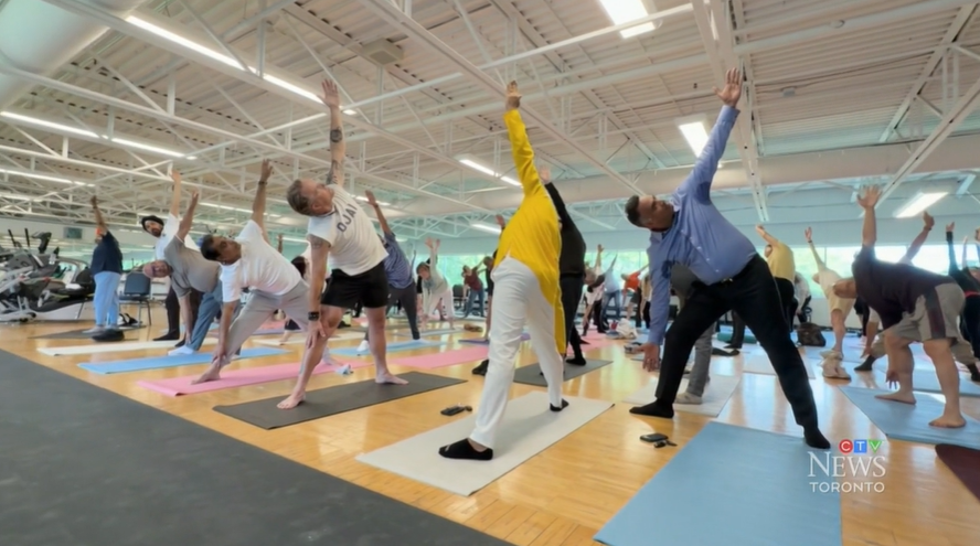 Participants in a group yoga class perform a standing side stretch on colourful yoga mats in a bright fitness studio, as part of a cardiac rehabilitation program at UHN.