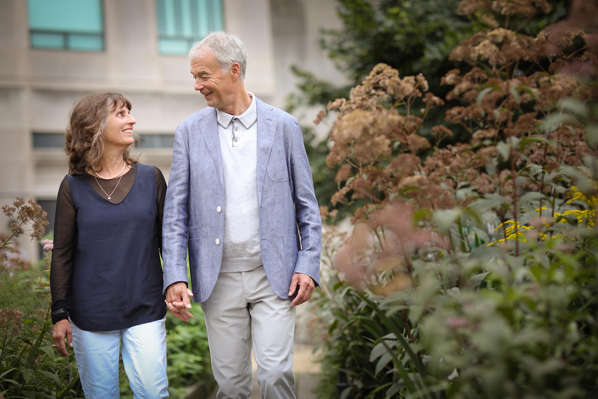 An older couple holding hands and smiling at each other while walking through a lush green garden outside a building.