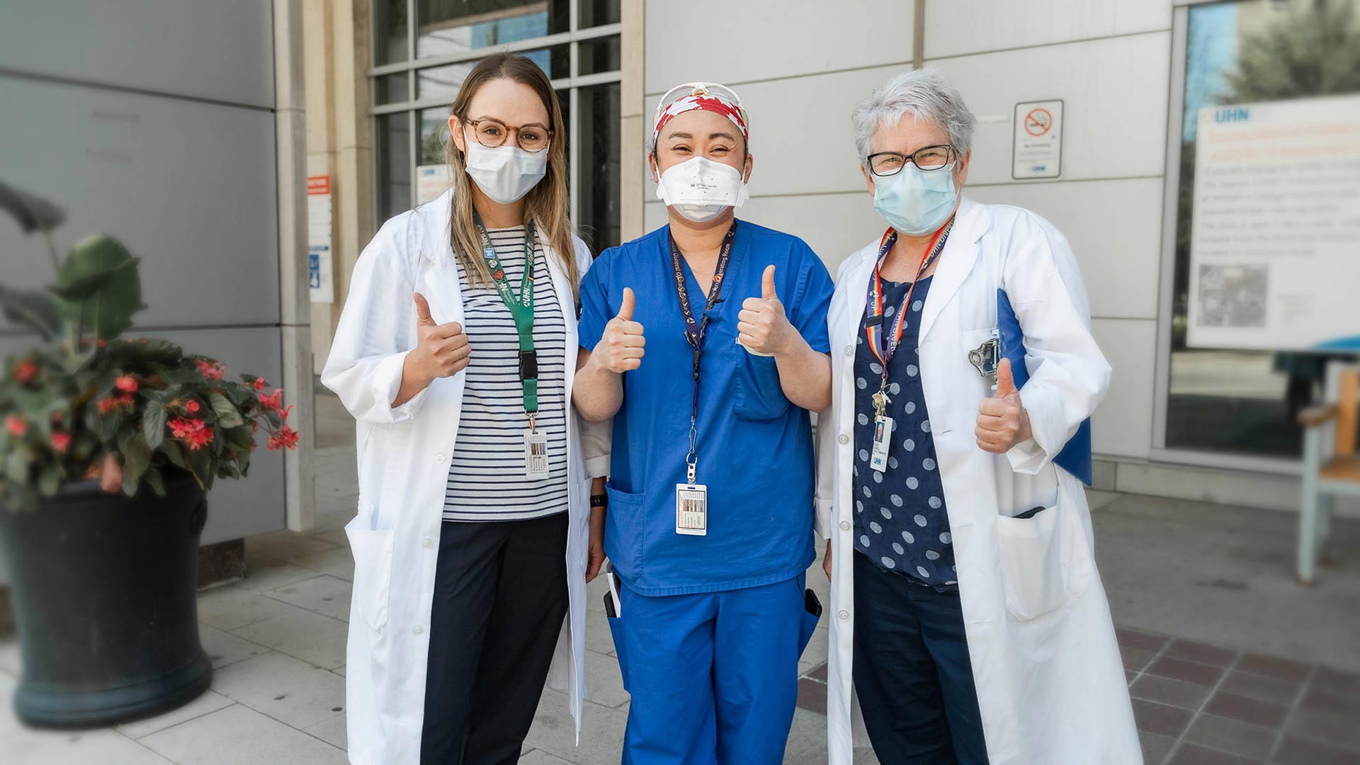 Three healthcare workers wearing face masks, white lab coats, and blue scrubs standing together outside a hospital building and giving a thumbs-up.