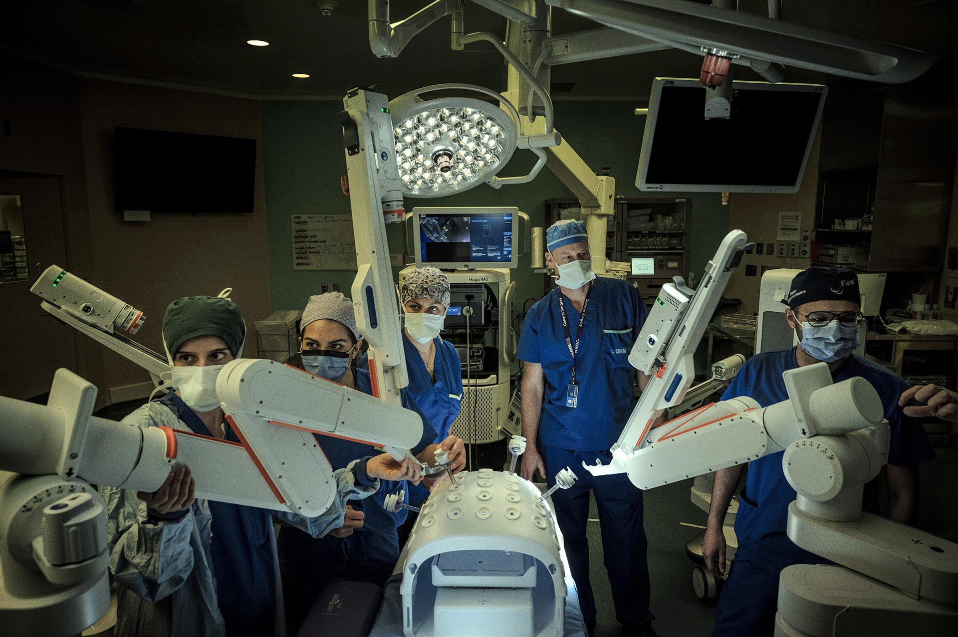 A team of medical professionals in scrubs and masks operating a HUGO robotic surgical system in an operating room.