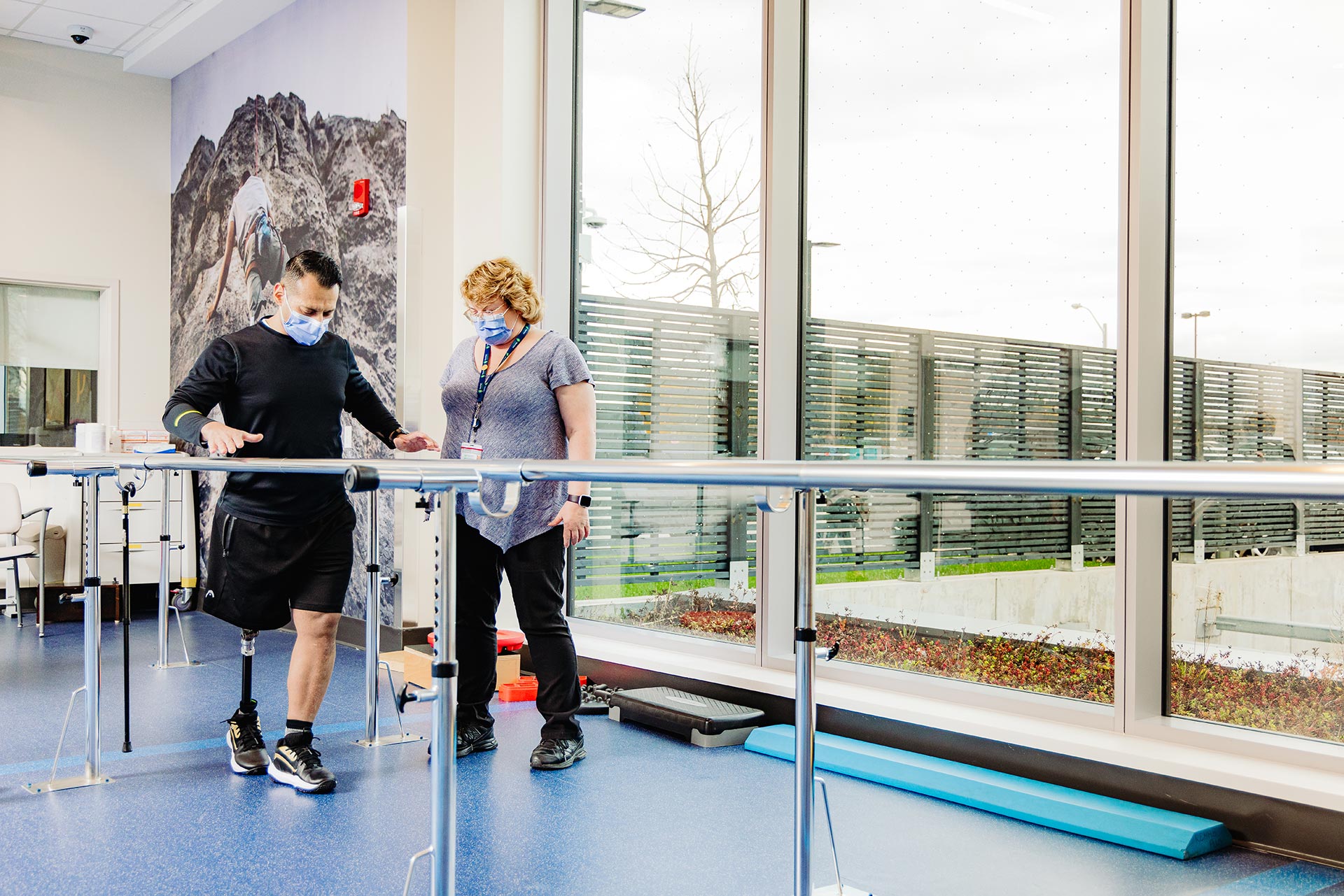 A physical therapist wearing a mask supervises a male patient with a prosthetic leg walking between parallel bars in a bright rehabilitation room.