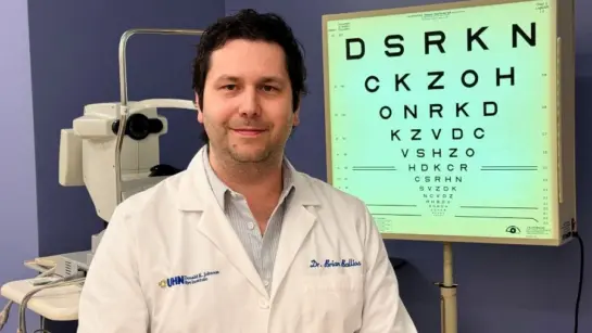 Dr. Brian Ballios sits in front of an eye chart in an eye examination room while wearing his white coat.