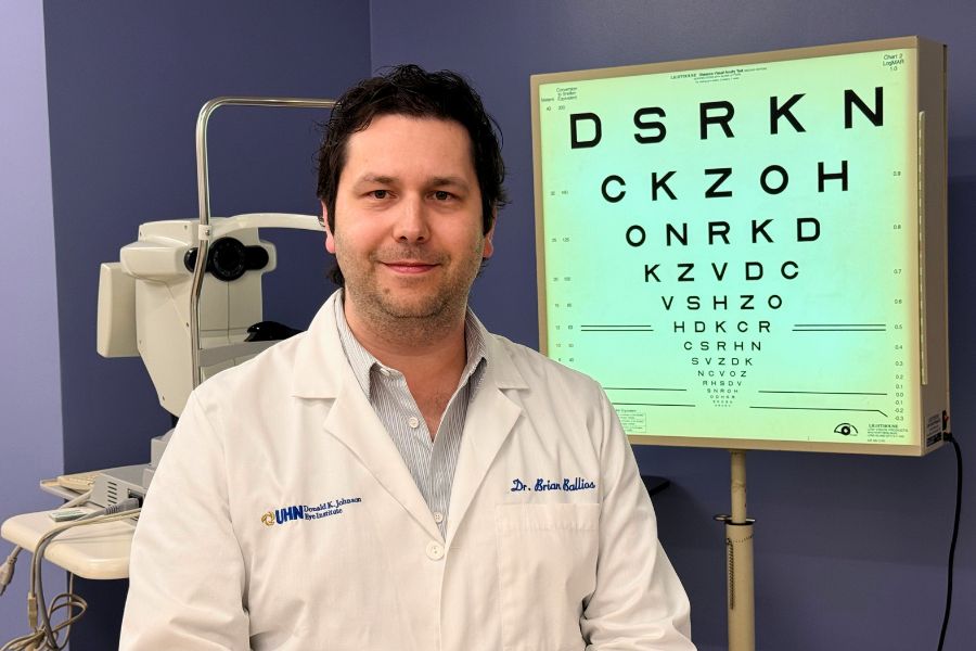 Dr. Brian Ballios sits in front of an eye chart in an eye examination room while wearing his white coat.
