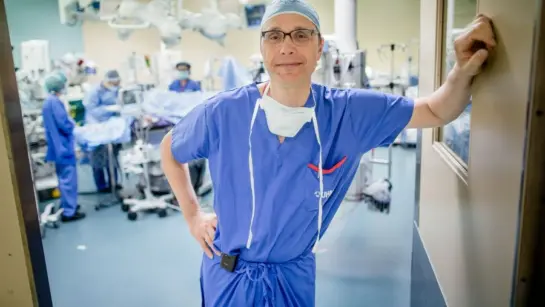 Dr. RJ Cusimano wearing his surgical scrubs and PPE while leaning against a doorway of an operating room and smiling for a photo.
