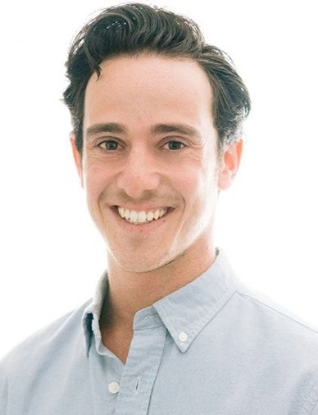 A headshot of Daniel Goldgut wearing a light blue dress shirt, smiling with his hair combed to the side with a bright white background.
