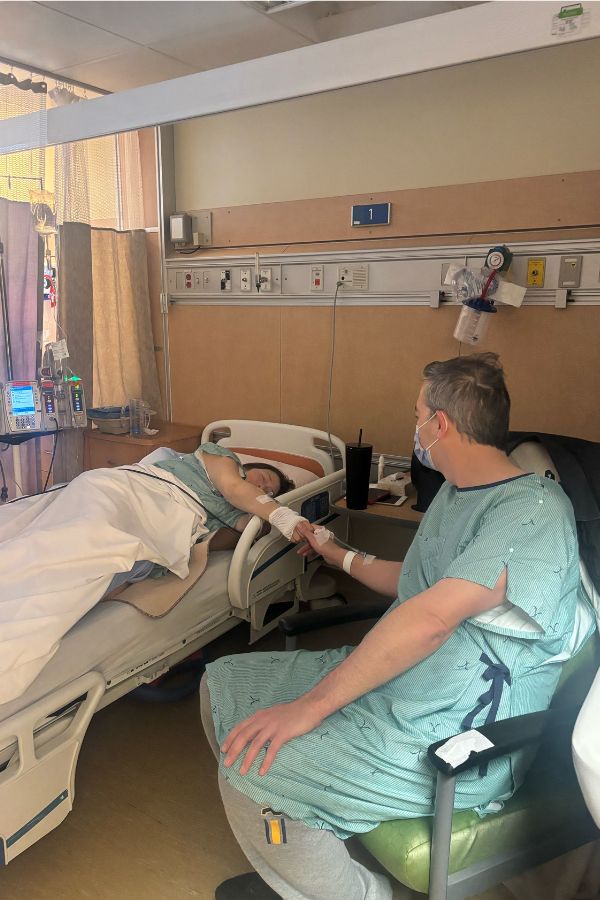 Mike seated next to his friend who's laying down in a hospital bed reaching out with her hand to Mike after receiving a transplant.
