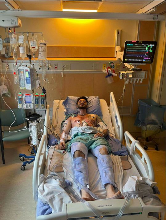 Joseph in a UHN hospital bed smiling while connected to various machines and devices.