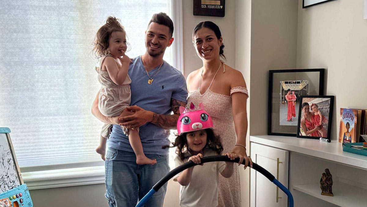 A smiling family of four poses indoors, with a father holding a toddler and a young child wearing a pink cat helmet standing in front of the mother.